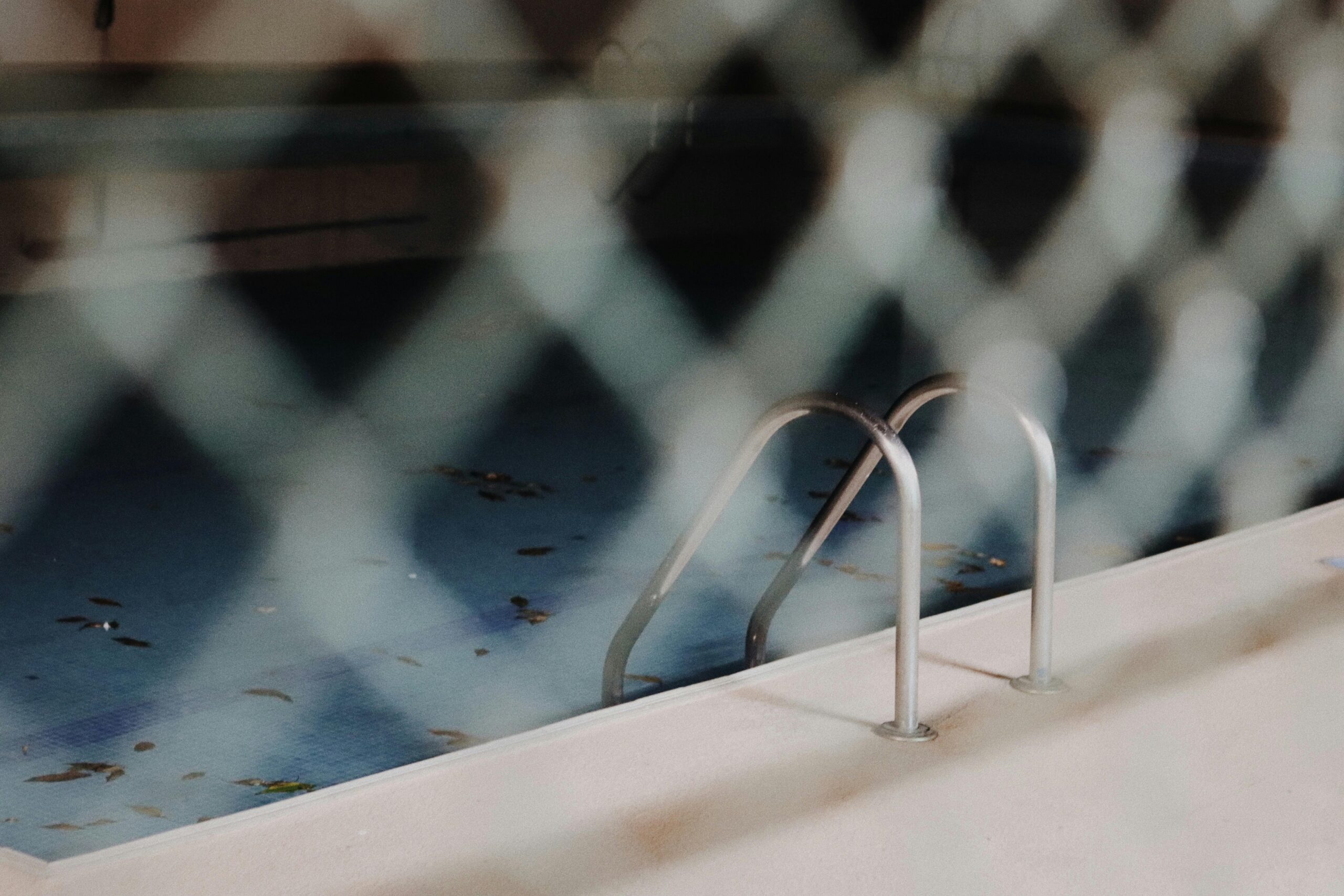 A view of a neglected swimming pool with a metal ladder, seen through a chain-link fence.