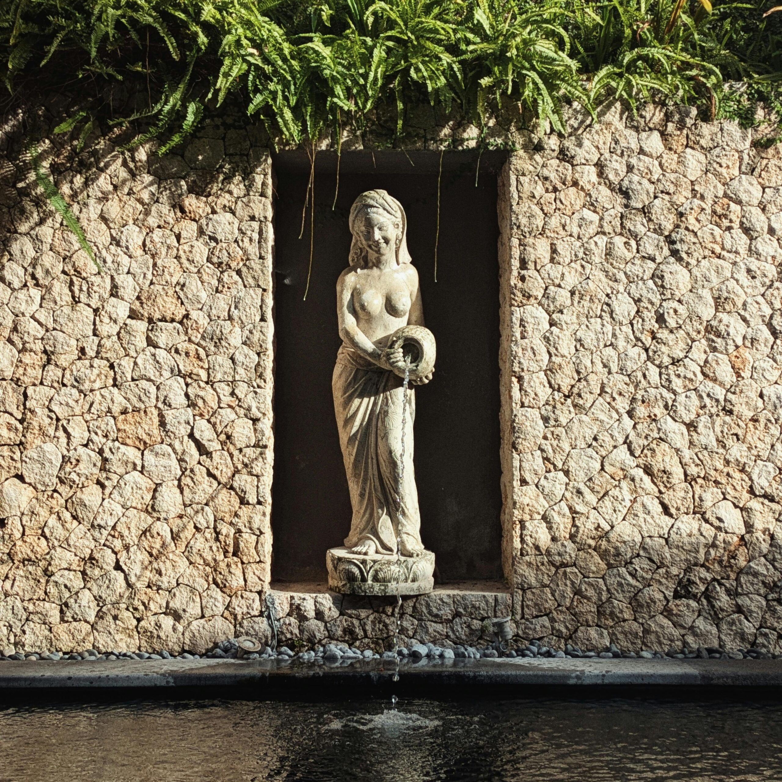 Stone statue of a woman in a garden alcove, surrounded by lush ferns and water feature.