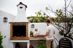 Man wearing casual clothes exploring outdoor kitchen with fireplace and sink together with cute toddler kid in backyard near white stone fence during daytime at countryside