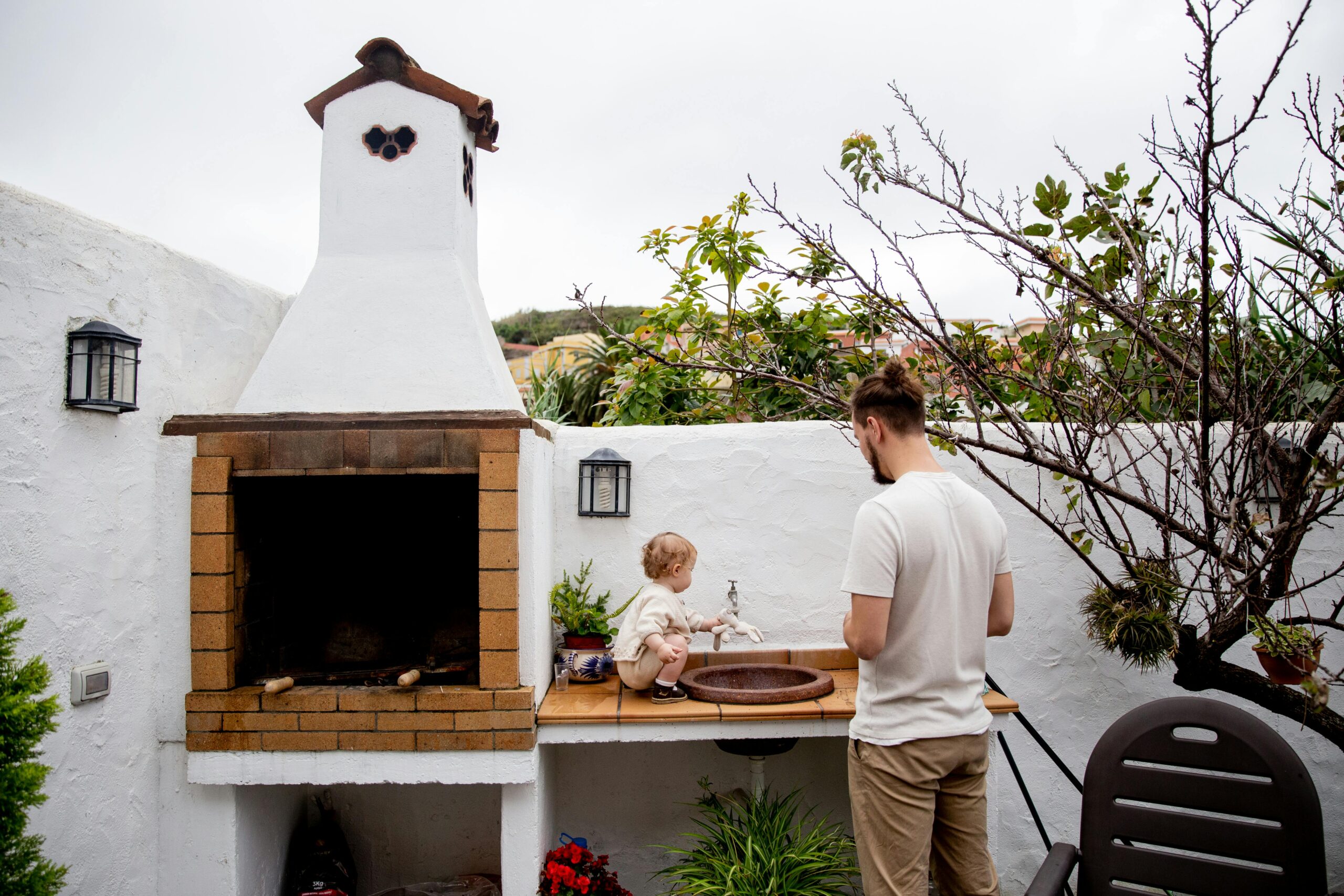 Man wearing casual clothes exploring outdoor kitchen with fireplace and sink together with cute toddler kid in backyard near white stone fence during daytime at countryside