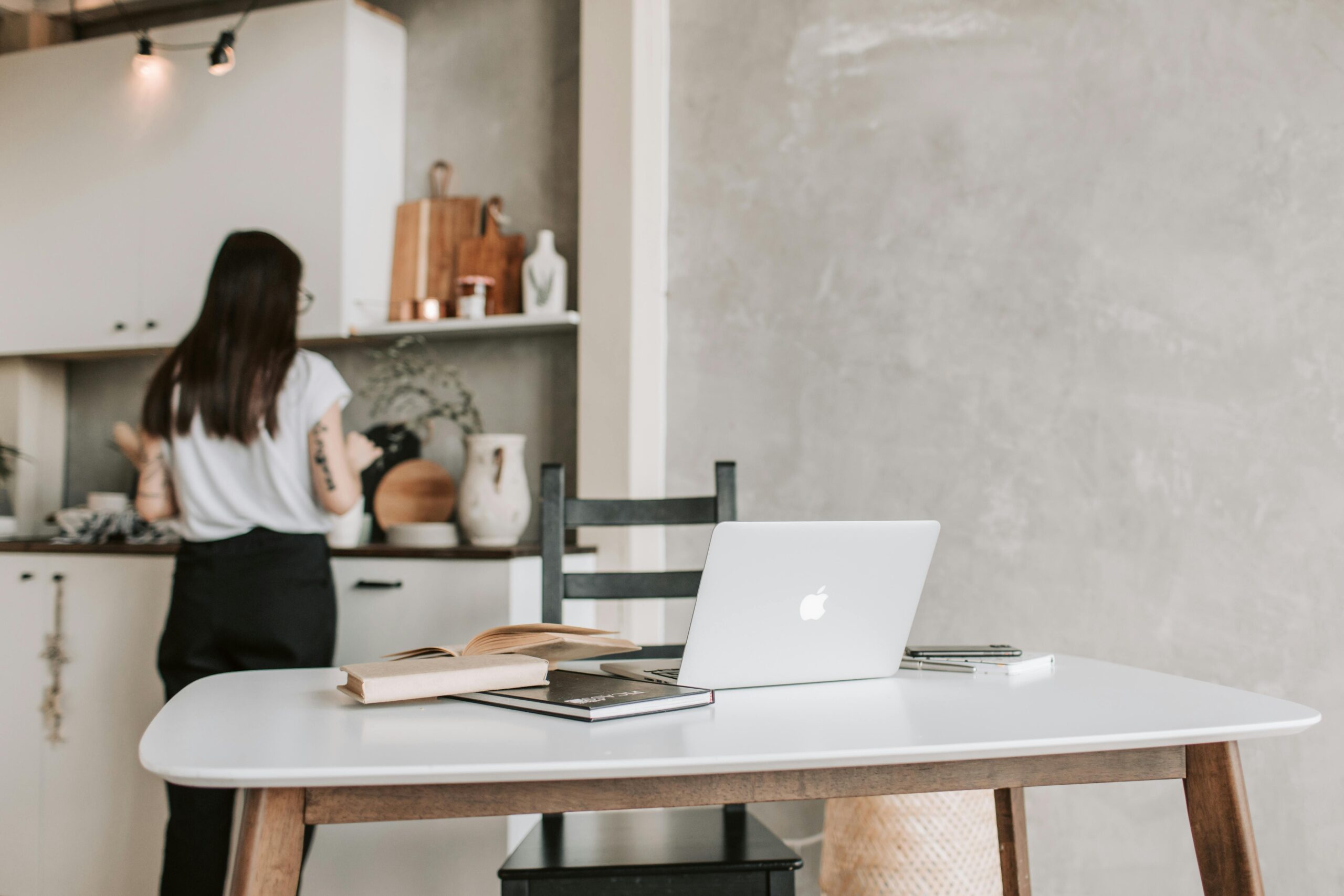 Laptop on a white table in a stylish home office, with a person in the kitchen.