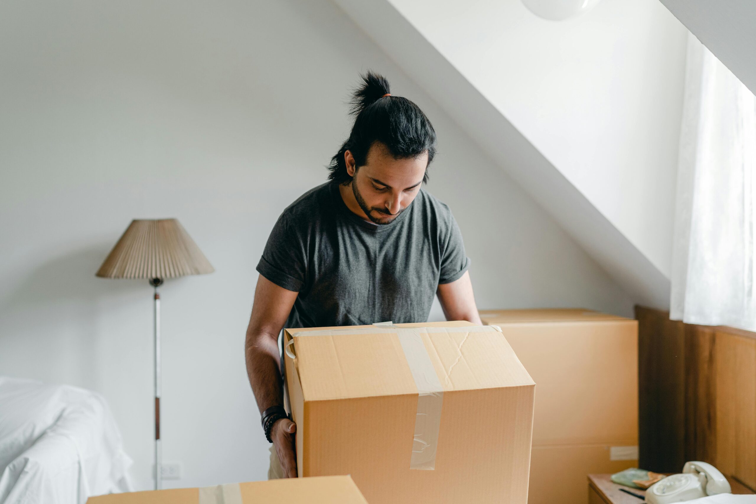 A man with a ponytail lifts a cardboard box in an attic room, suggesting relocation.