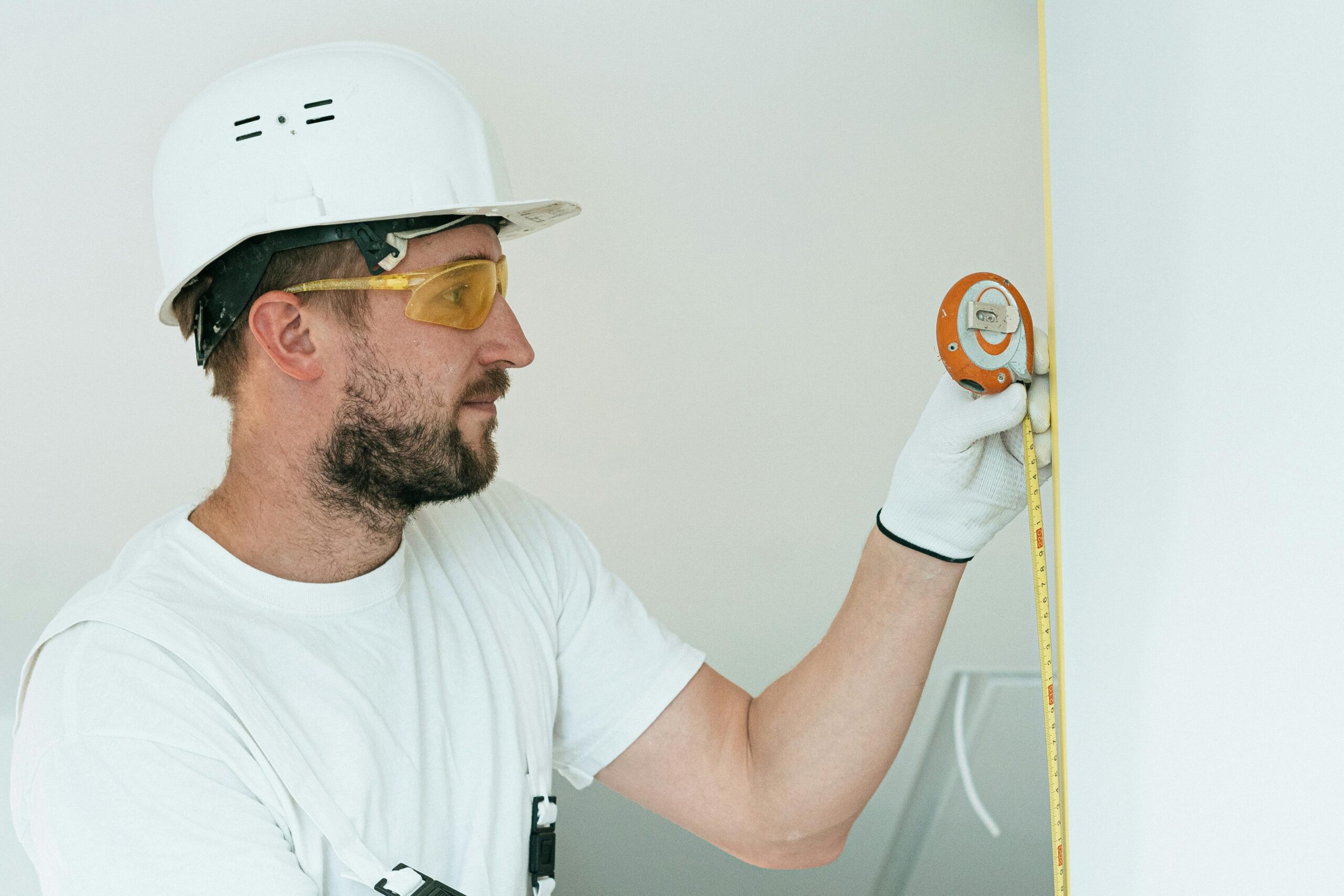 Construction worker with hard hat and goggles measuring a wall with a tape measure.