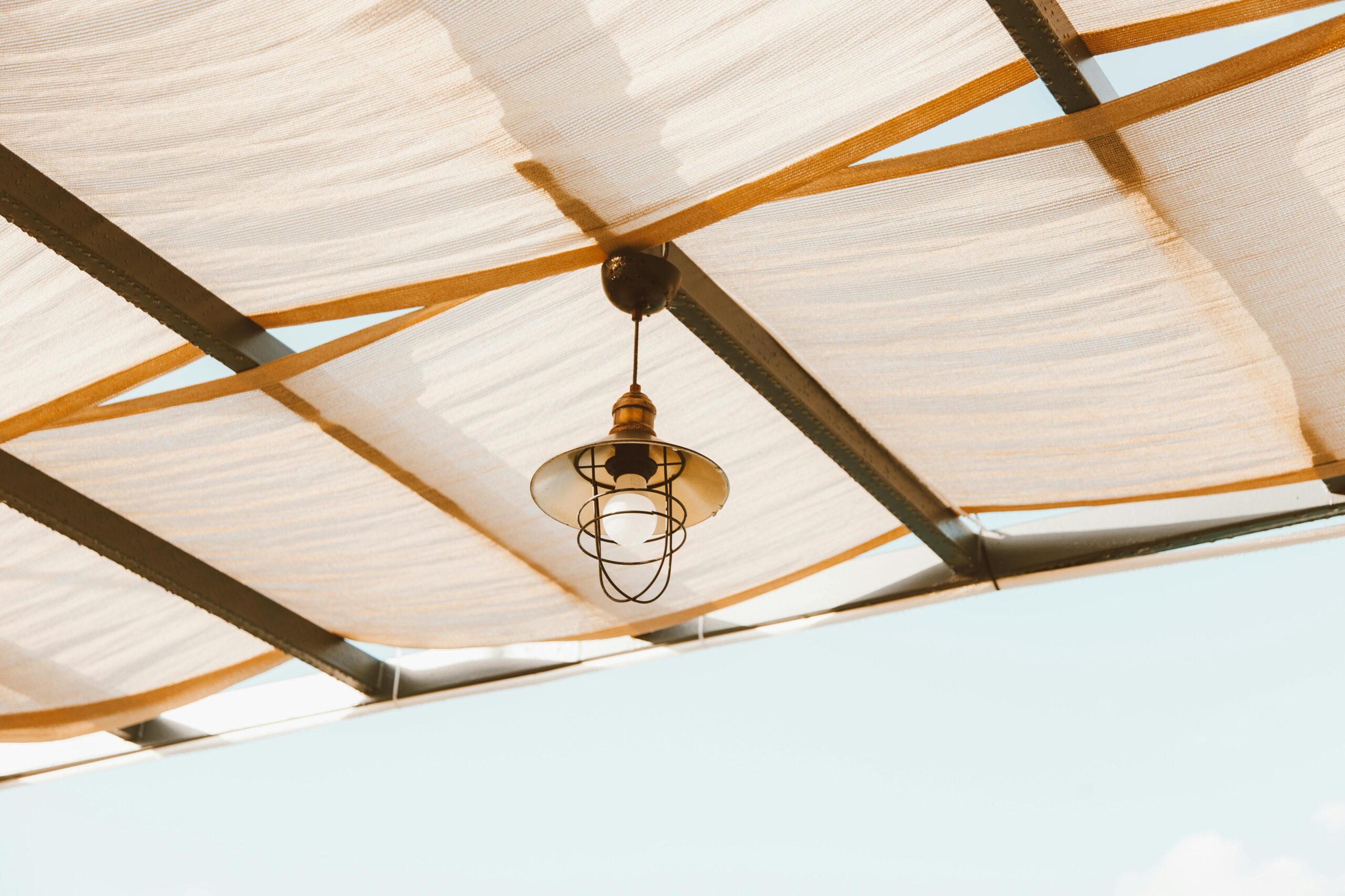 Minimalist industrial lamp hanging under a fabric ceiling against a clear blue sky.