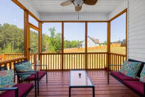 An inviting enclosed porch with cozy furniture overlooking lush greenery on a sunny day.