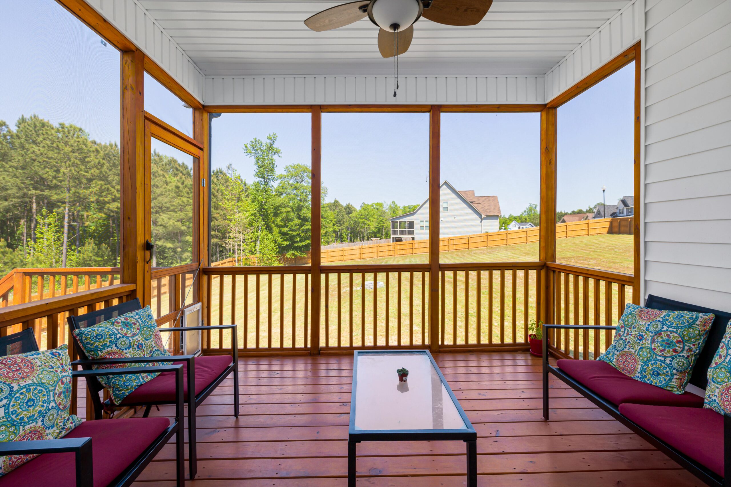 An inviting enclosed porch with cozy furniture overlooking lush greenery on a sunny day.